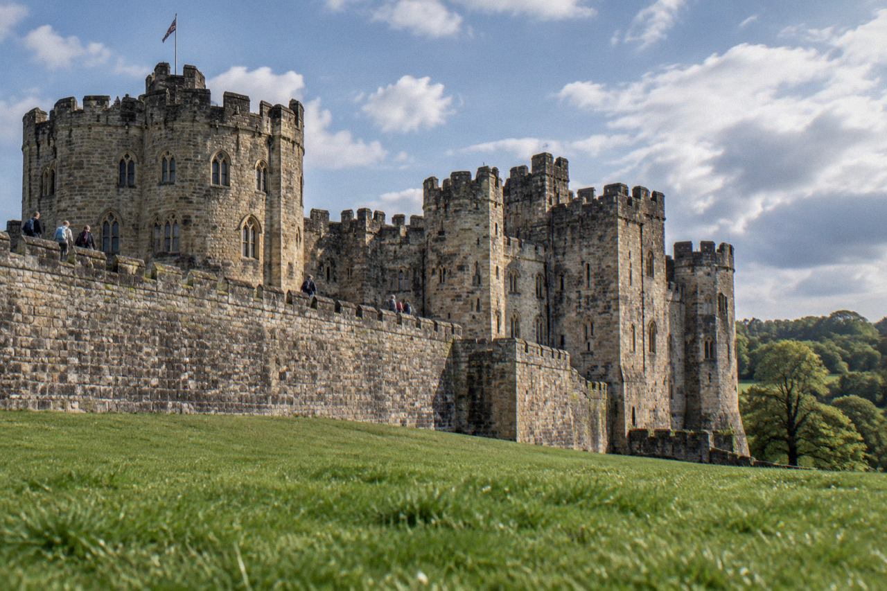 Low angle of Alnwick Castle's stone towers and battlements from the grassy Outer Bailey.