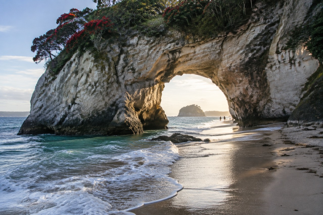 Large natural rock arch over white sand beach with turquoise water and flowering trees