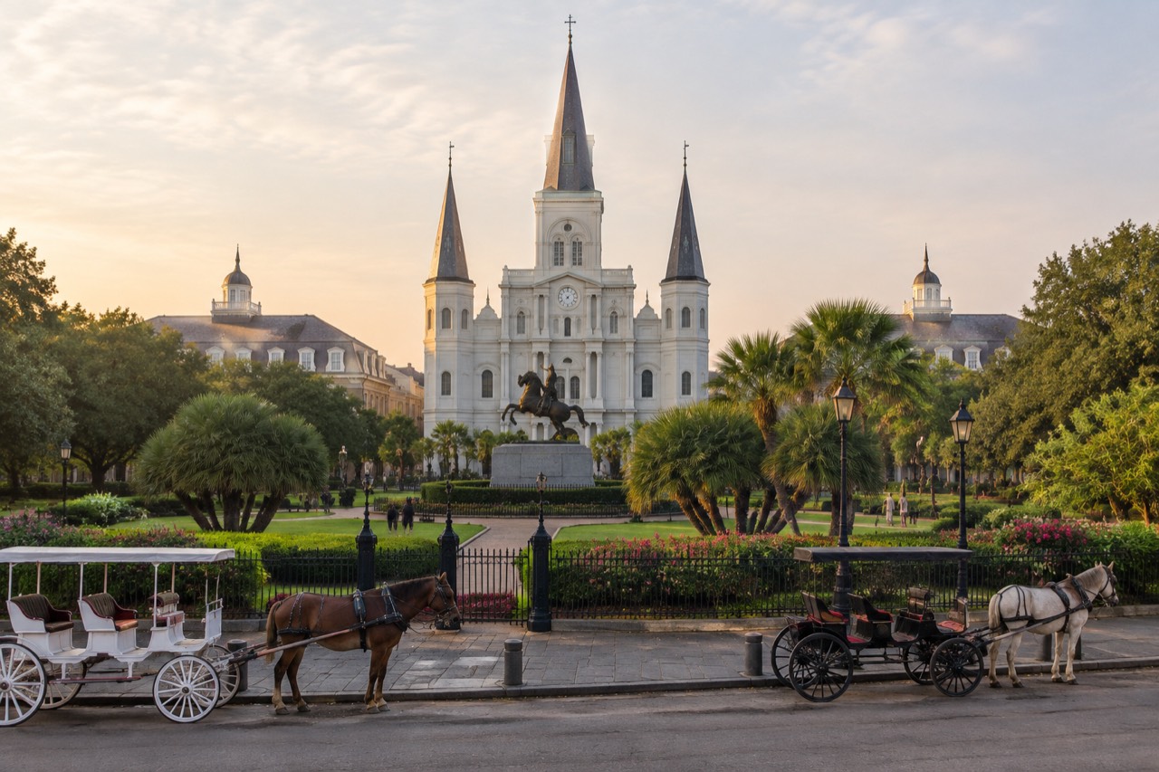 Sunlit cathedral twin spires behind a fenced garden square with a bronze horse statue