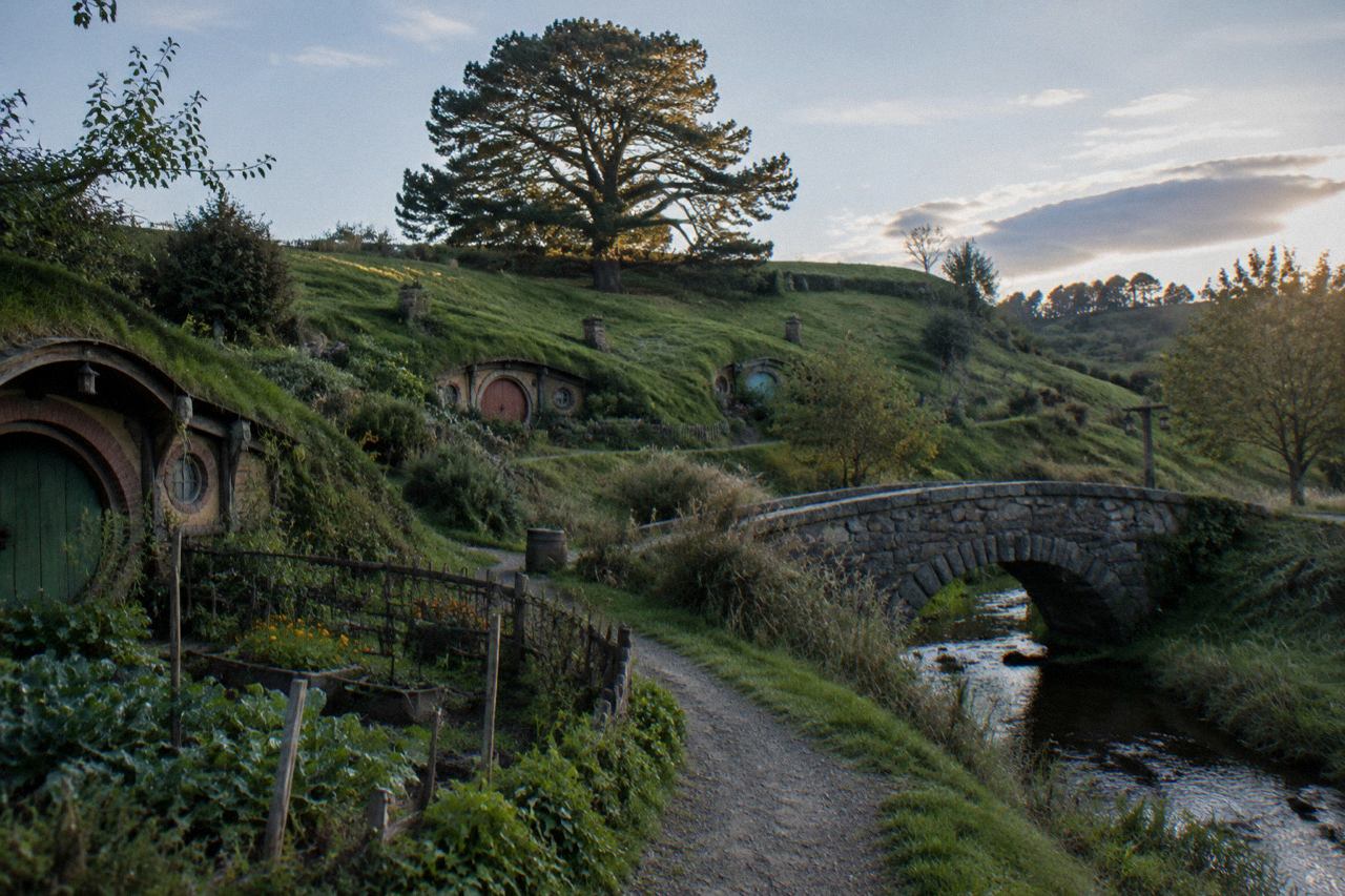 Round-doored cottages tucked into green hills beside a lone tree, golden afternoon light.