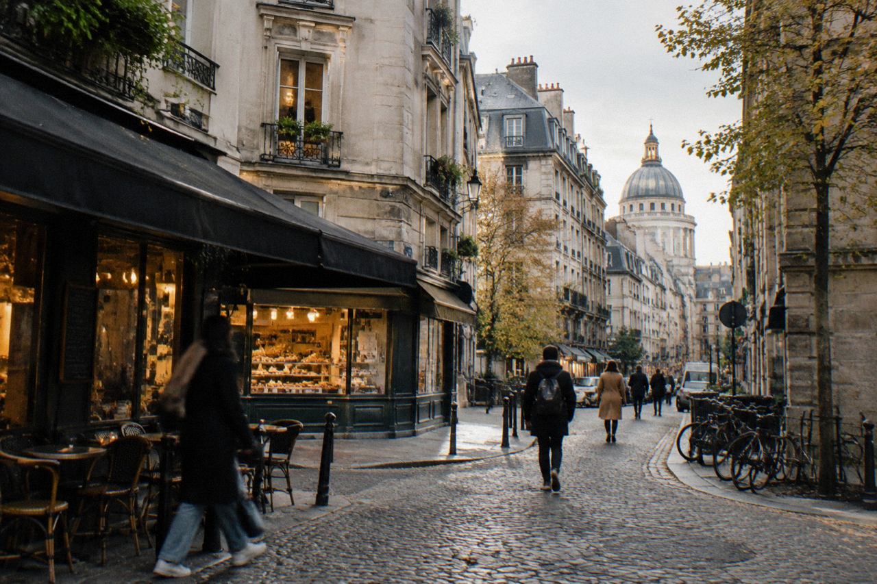 Cobbled Paris square at morning with bistro awning, bakery windows, and Panthéon dome above rooftops.