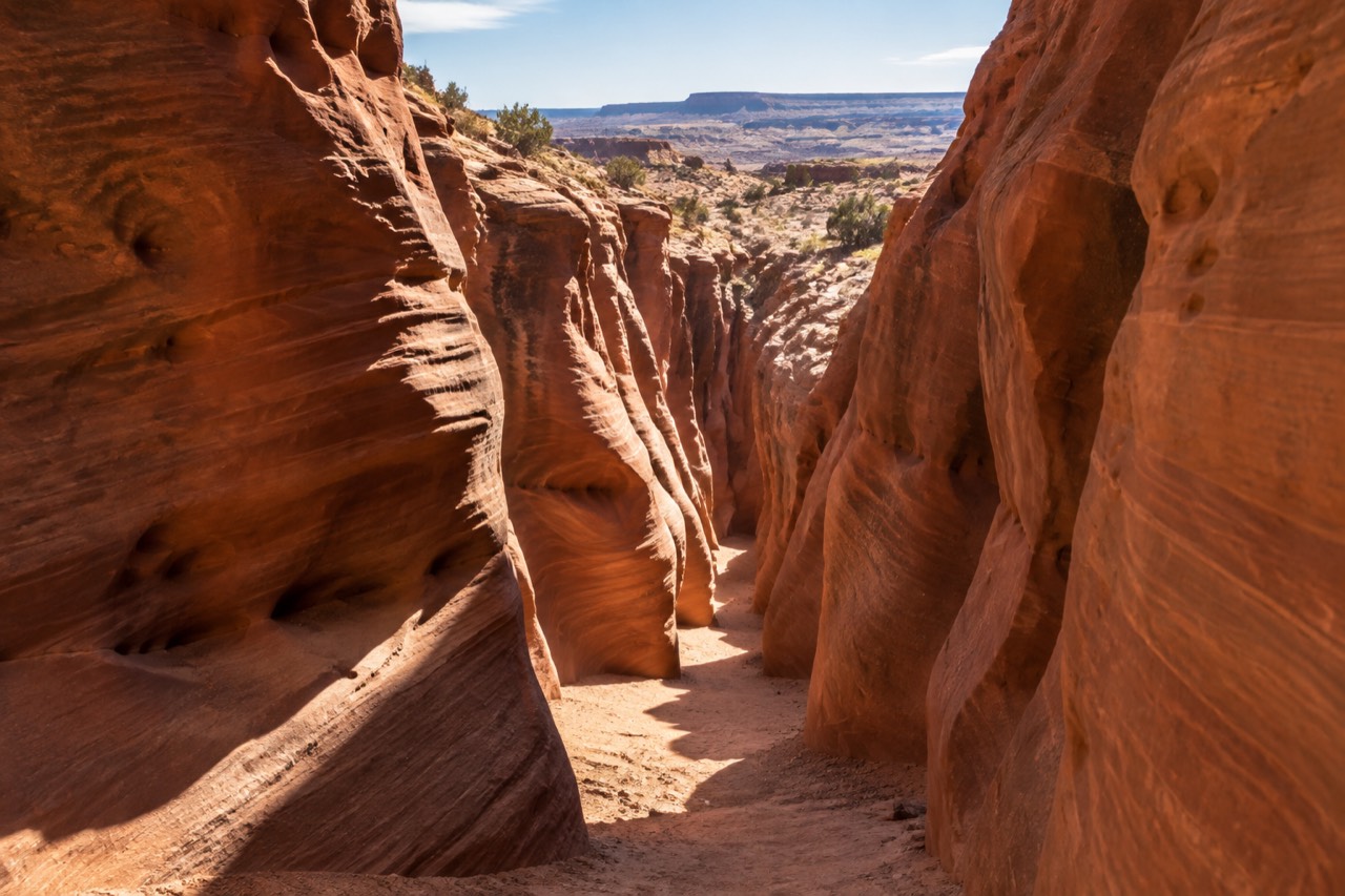 Sunlight beams into a narrow red sandstone slot canyon with a sandy floor.