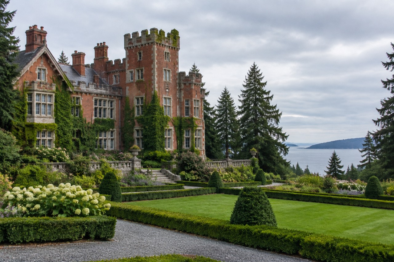 Red-brick castle with turrets behind formal gardens and tall firs on an overcast morning.