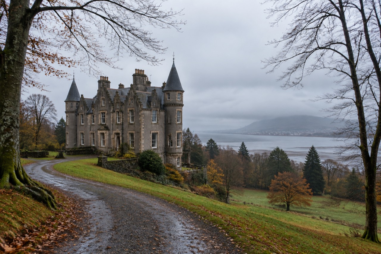 Grey stone turreted mansion on a wooded hill above Dumbarton, Clyde estuary beyond, overcast morning.