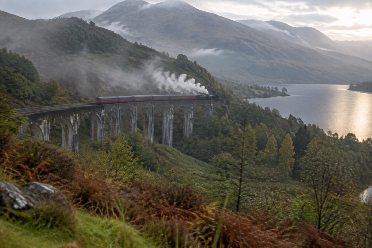 Steam train crossing stone viaduct arches above a green Scottish glen and loch