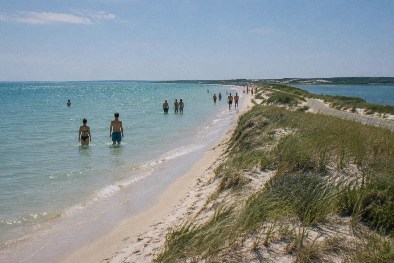 Calm shallow beach, grassy dunes and distant swimmers under a hazy summer sky.