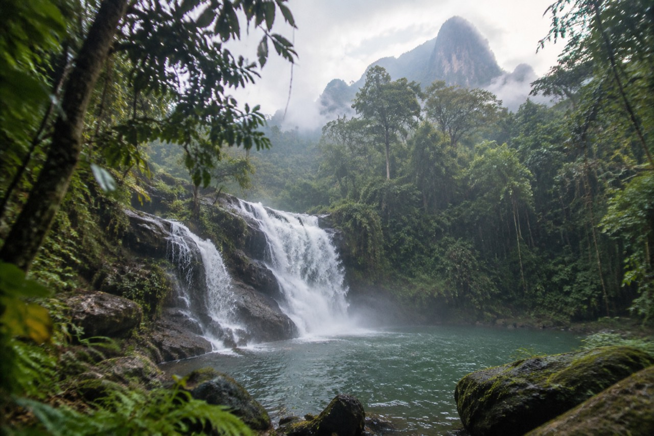 Misty rainforest waterfall cascading over dark limestone into a green pool in Krabi, Thailand