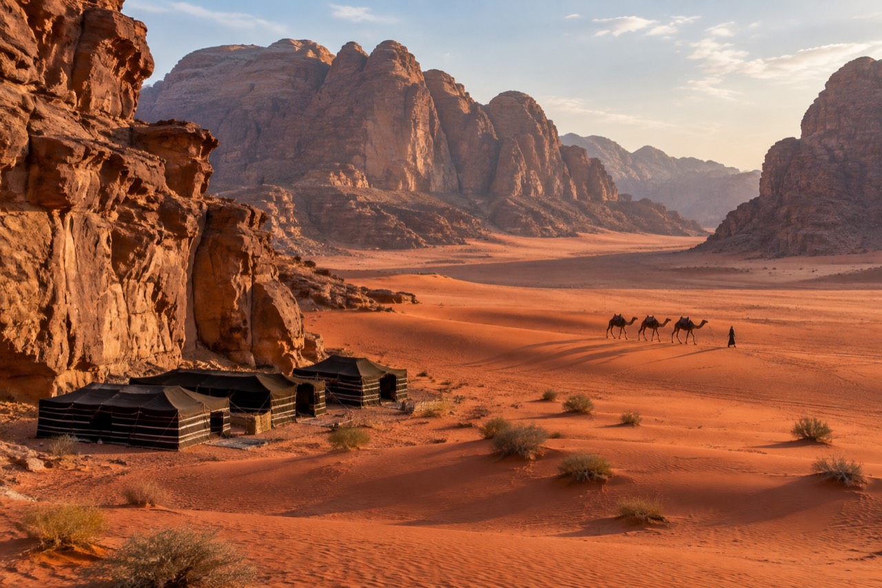 Red desert valley with towering sandstone cliffs, black Bedouin tents, and distant camel caravan at sunset.