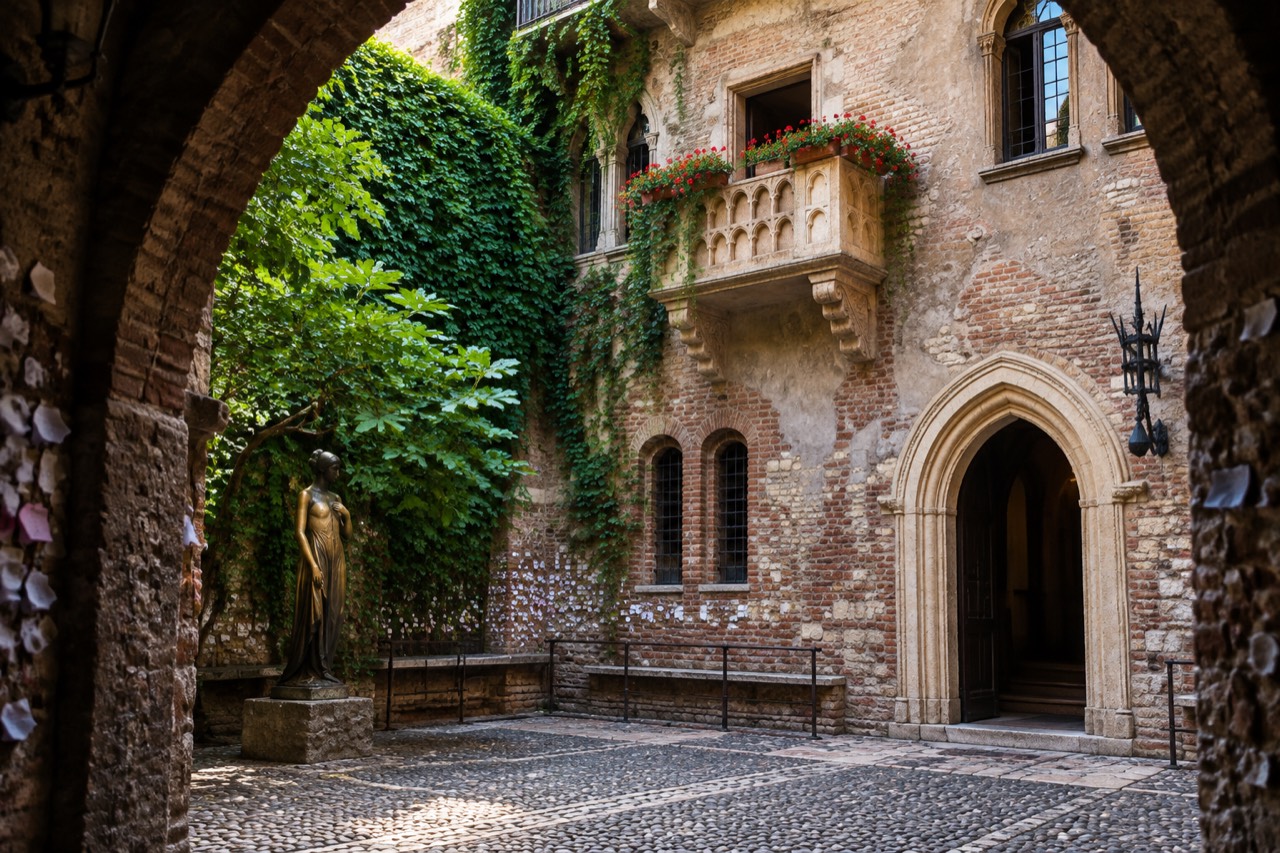 Sunlit Verona courtyard with stone balcony, geraniums, paper notes in walls, bronze statue