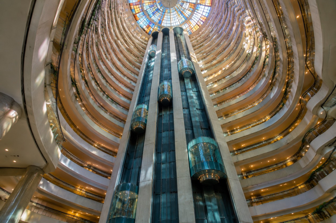 Glass elevators climbing a huge curved hotel atrium beneath a colorful skylight