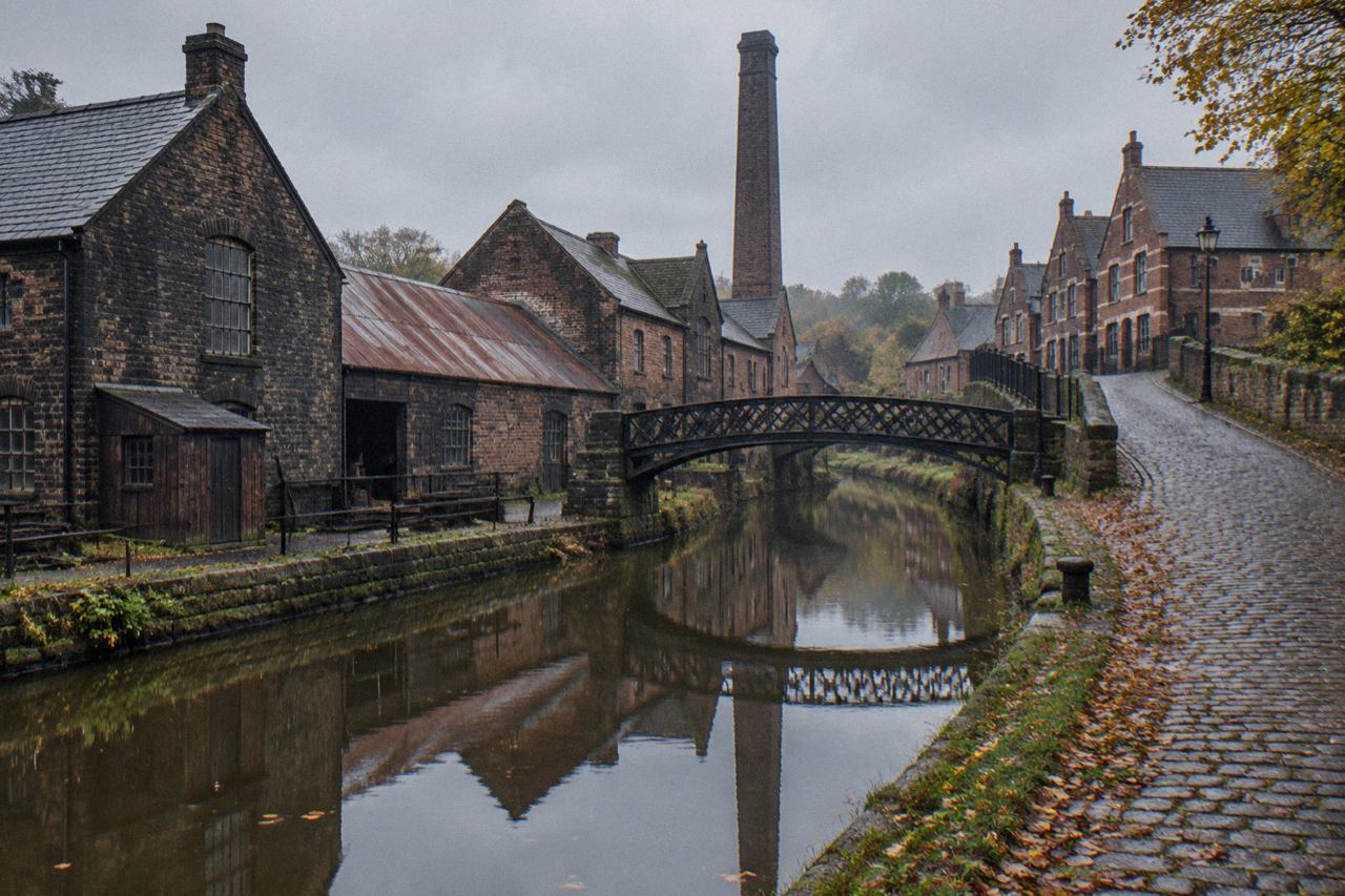 Cobbled canal-side street with dark brick workshops, iron footbridge and chimney at a Victorian industrial museum.