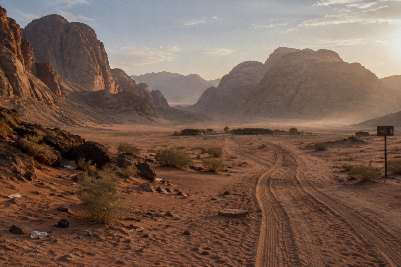 Red desert valley with towering sandstone cliffs, long shadows, and distant tents under hazy sky.
