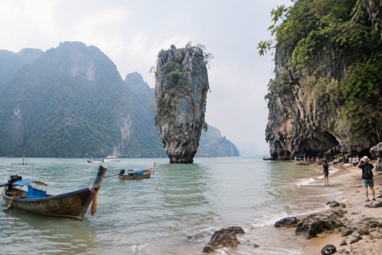 Limestone pinnacle in emerald bay beneath a leaning cliff, longtail boats and pale beach nearby.