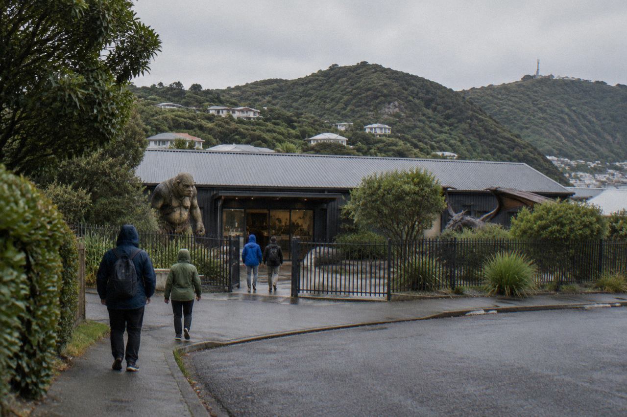 Low industrial workshop building on a leafy suburban street beneath green hills under grey sky.