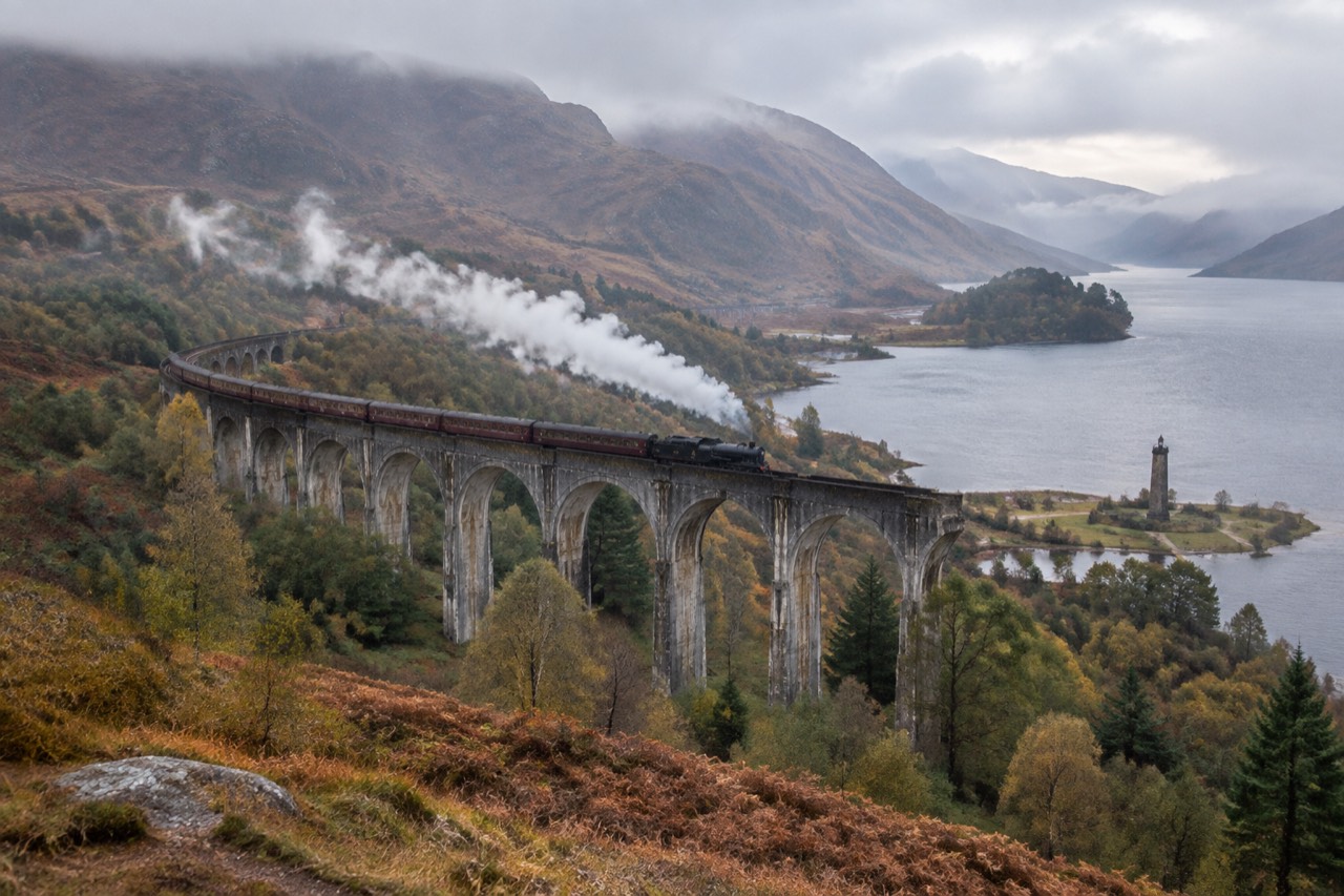 Steam train crossing curved Glenfinnan viaduct above Loch Shiel, stone monument tower, autumn morning mist