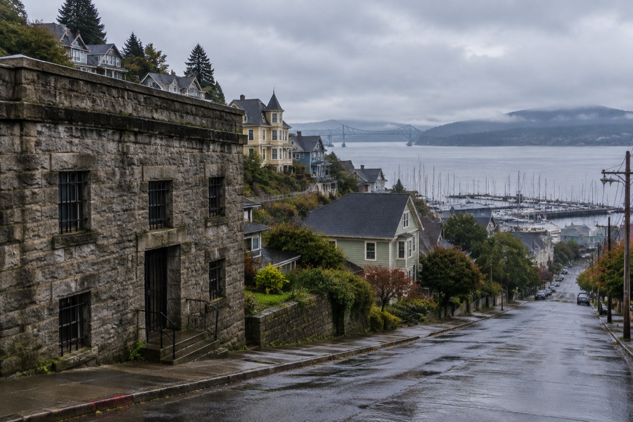 Stone jail building with barred windows beneath Astoria's hillside of Victorian houses.