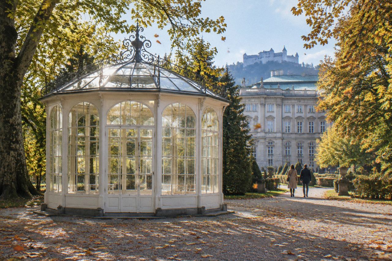 Glass gazebo with iron cresting in a palace park, autumn sun, distant visitors